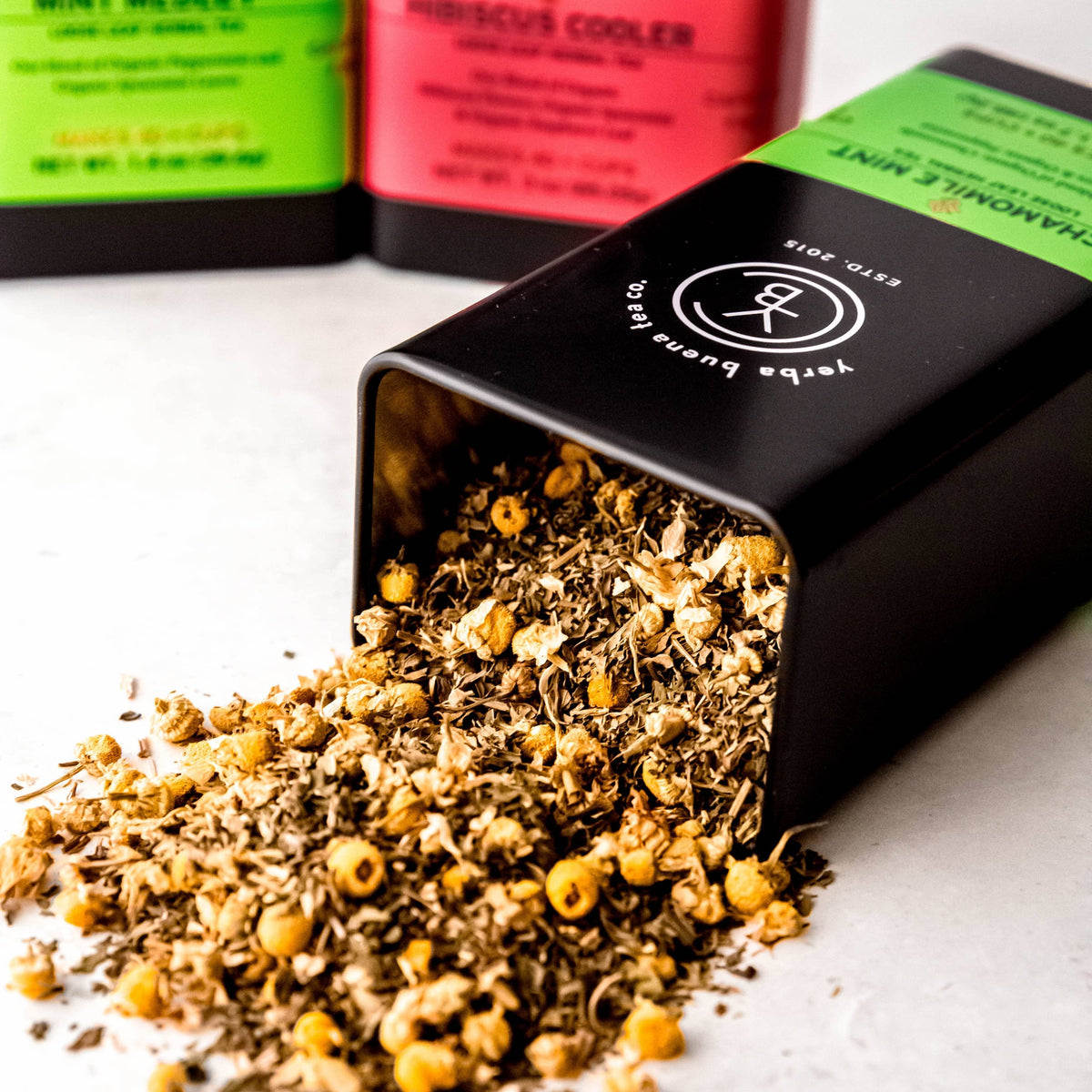 Loose leaf herbal tea spilling from a chamomile mint tin beside colorful gift tins, by Yerba Buena Tea Co.