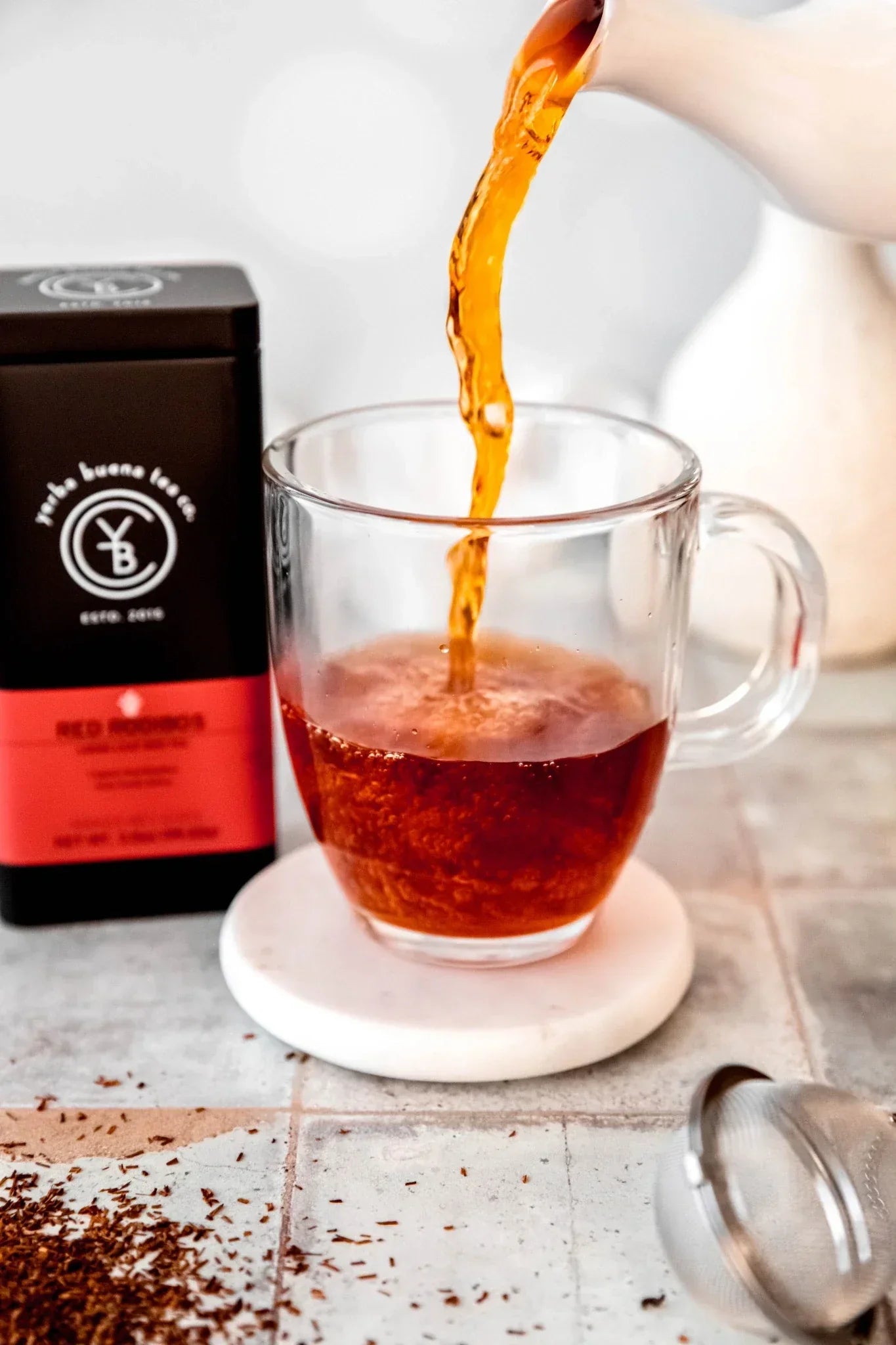 A rich amber cup of organic red rooibos tea being poured into a glass mug, with loose red tea leaves and a tea infuser, by Yerba Buena Tea Co.
