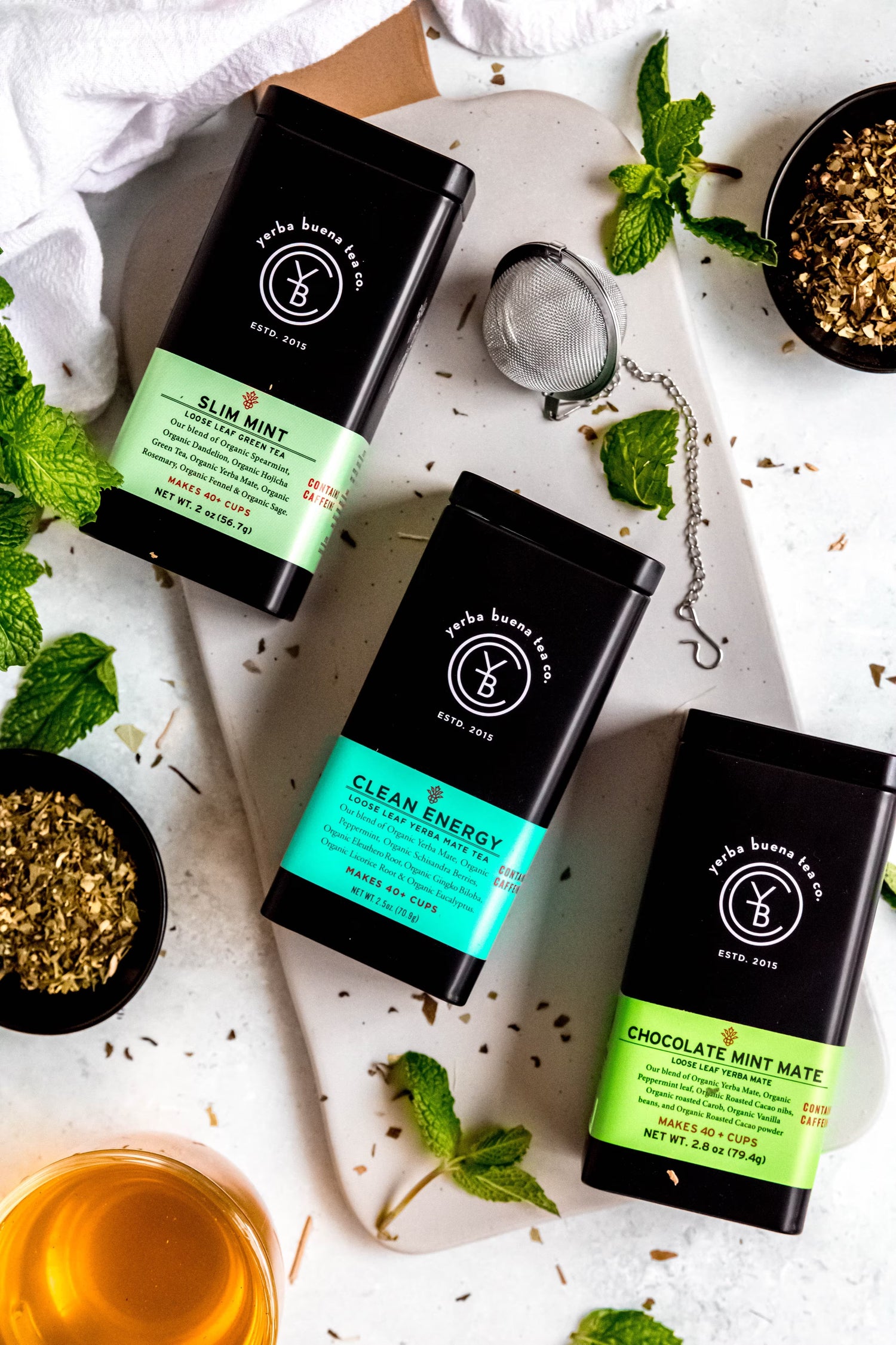 Flatlay of Yerba Buena Tea Co. organic yerba mate tins (Slim Mint, Clean Energy, Chocolate Mint Mate) with fresh mint leaves and a glass of brewed tea.