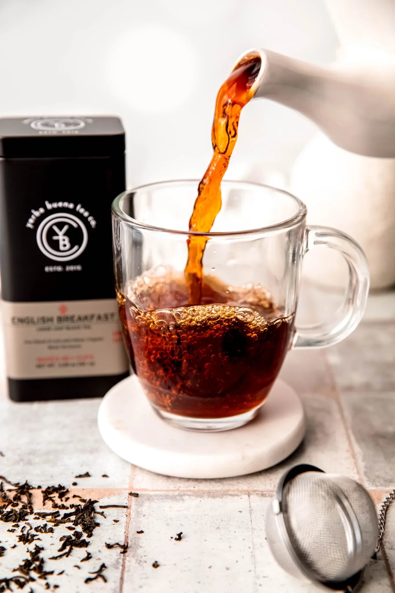 English Breakfast tea steaming into a clear glass mug with loose black tea leaves scattered on a white surface and branded black tin container nearby, by Yerba Buena Tea Company.