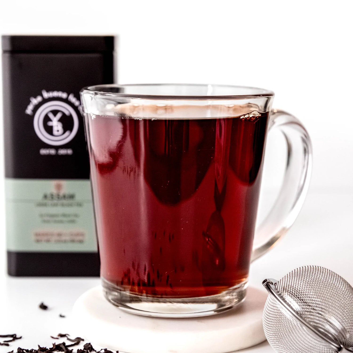 Brewed dark red Assam tea in clear glass mug with black pouch packaging behind it, by Yerba Buena Tea Company.