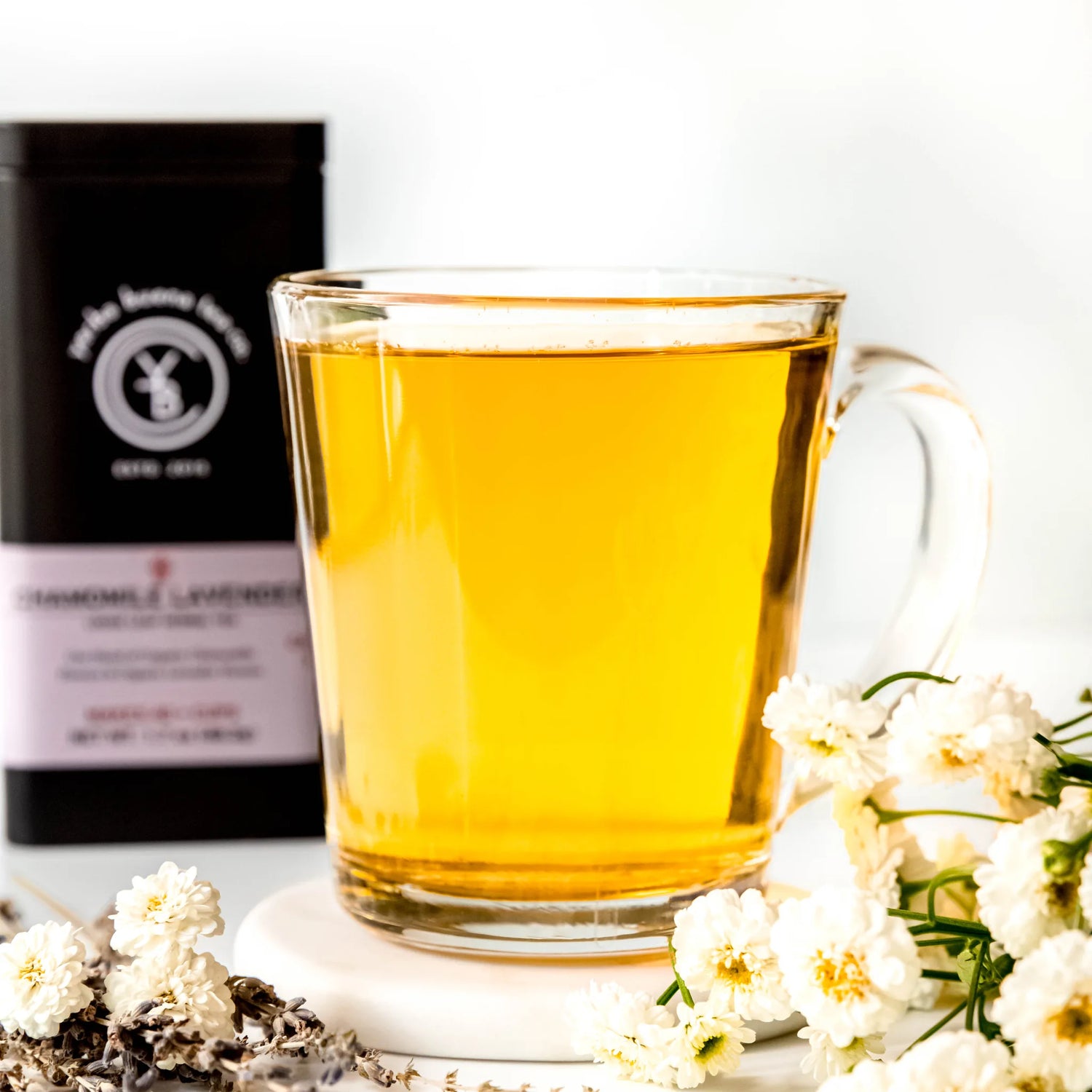 Brewed golden chamomile tea in clear glass mug surrounded by white flowers and loose leaves, by Yerba Buena Tea Company.