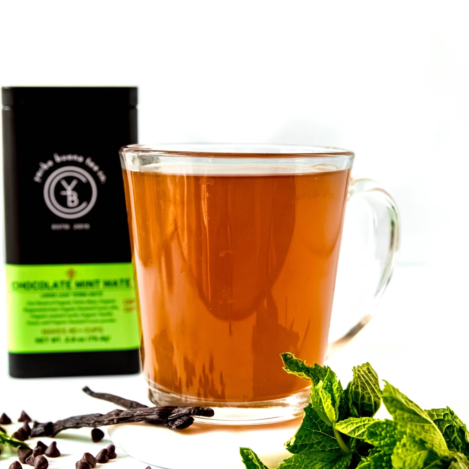 Brewed chocolate mint mate tea in a clear glass mug beside a black pouch with green label and fresh mint leaves, by Yerba Buena Tea Company.