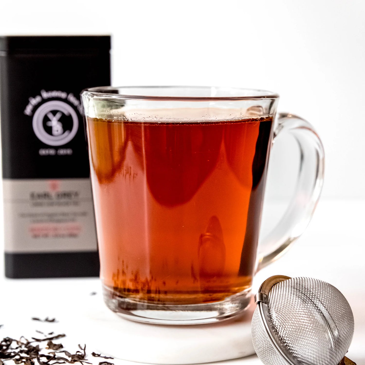 Deep red brewed Earl Grey tea in clear glass mug with infuser ball and black packaging box, by Yerba Buena Tea Company.