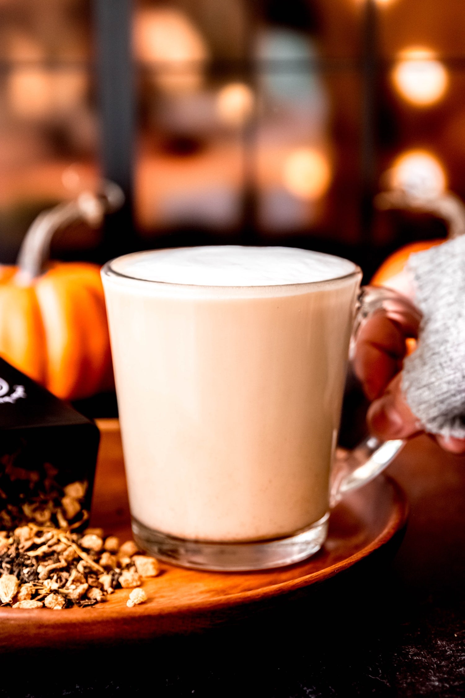 A hand in a cozy sweater holding a frothy chai latte in a glass mug, with pumpkins and warm lights in the background