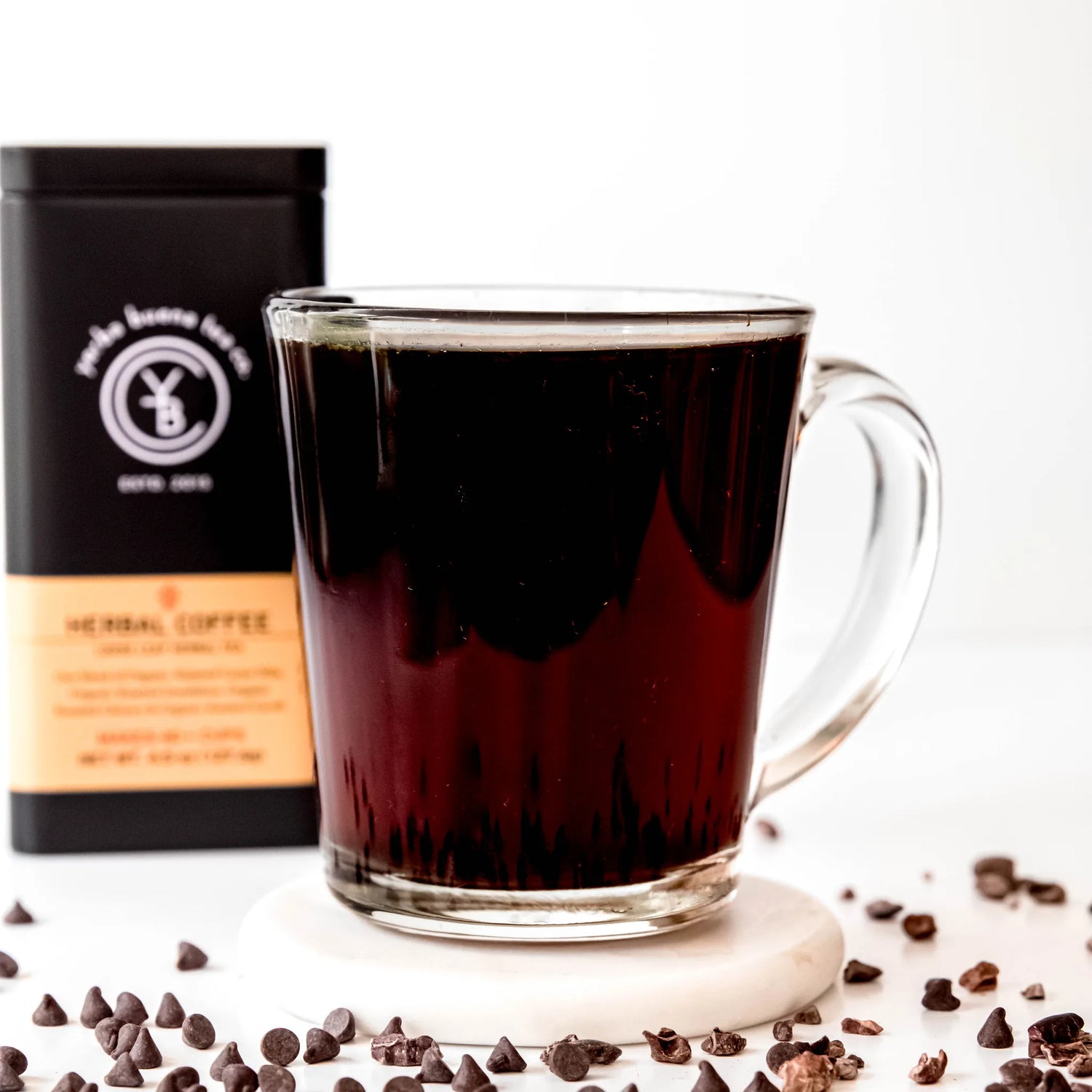 Dark brewed herbal coffee in a clear glass mug surrounded by scattered coffee beans and a black pouch, by Yerba Buena Tea Company.