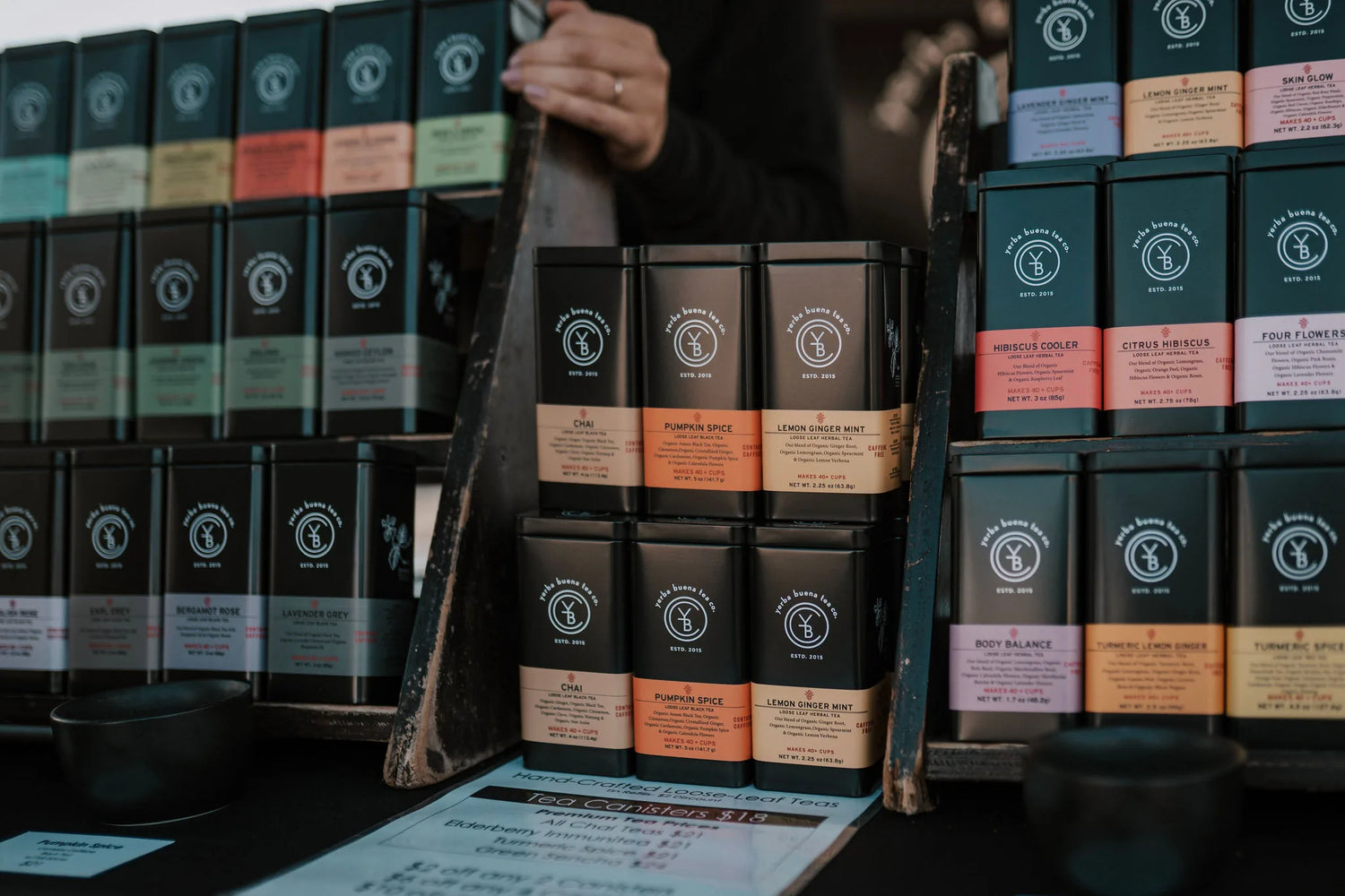 Tea booth display featuring organized black canisters with Chai, Pumpkin Spice, and Lemon Ginger Mint varieties, with a vendor's hand visible and product shelving in background, Yerba Buena Tea Company.