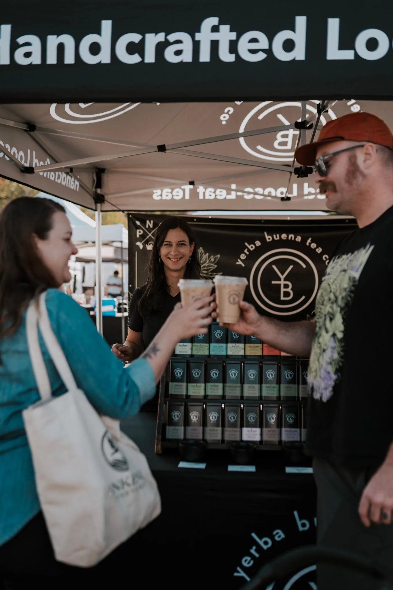 Vendor and customer clinking tea sample cups at a pop-up booth with organized tea tin display and branded signage visible, Yerba Buena Tea Company.