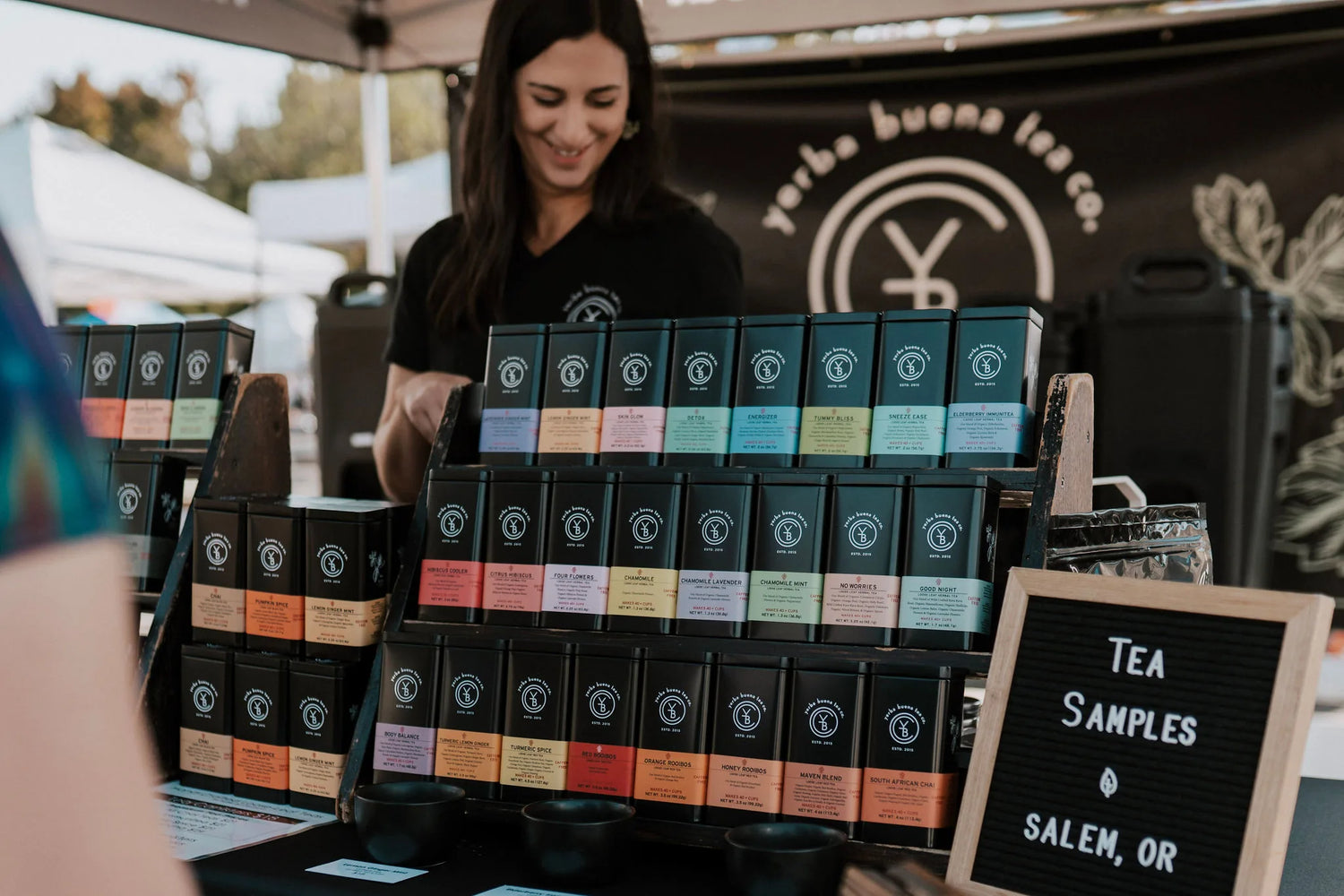 Vendor arranging tiered display of colorful tea boxes at outdoor booth with sampling station and branded signage visible, Yerba Buena Tea Company.