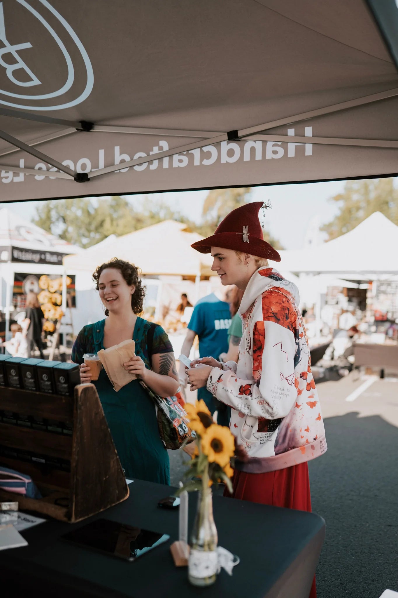 Vendor handing a paper bag to a smiling customer at a tea booth under a branded canopy at an outdoor farmers market, Yerba Buena Tea Company.