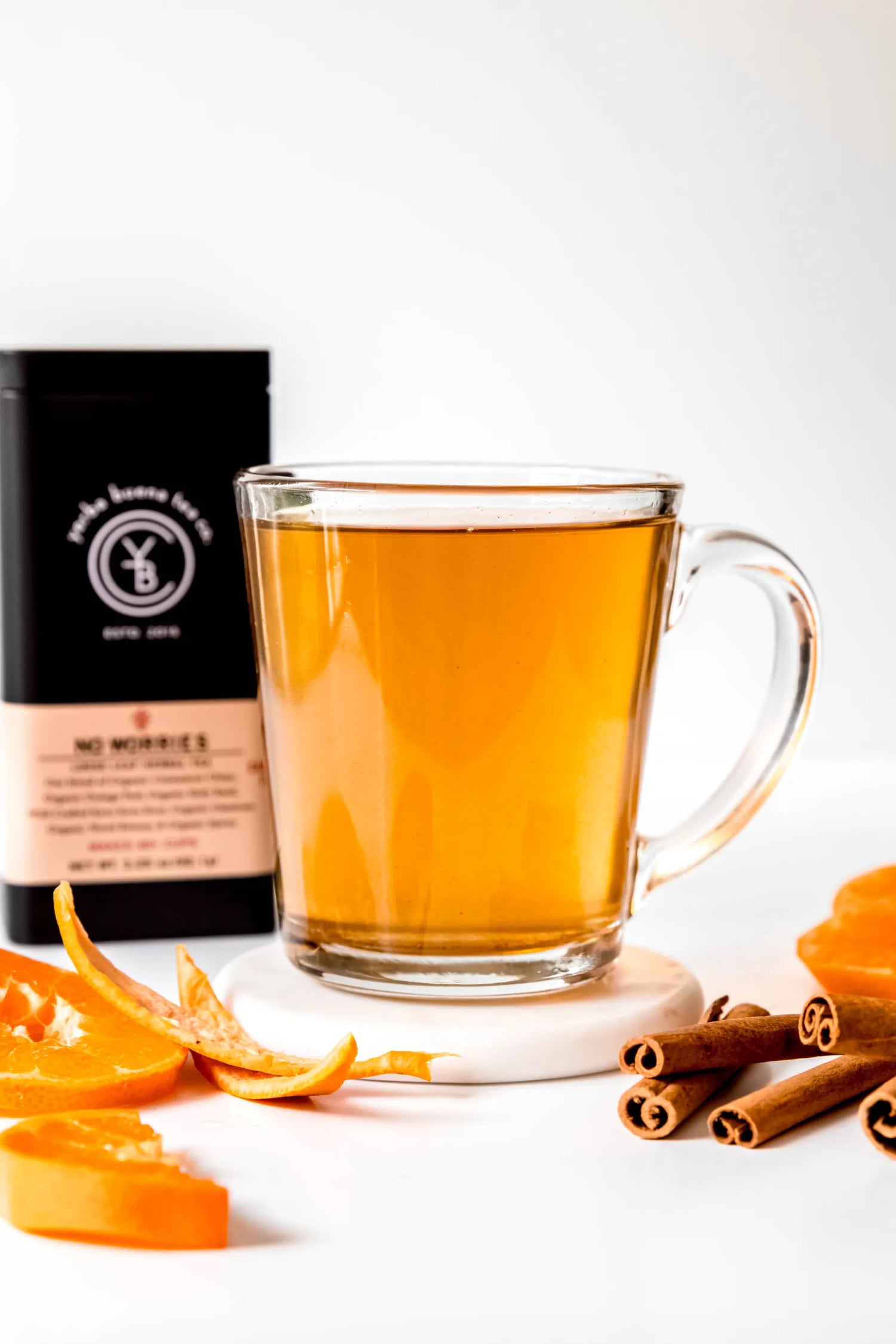 Clear glass mug filled with brewed No Worries organic herbal tea, warm amber in color, resting on a white surface with cinnamon sticks and orange peel nearby. A calming, caffeine-free tea ritual handcrafted by Yerba Buena Tea Co.