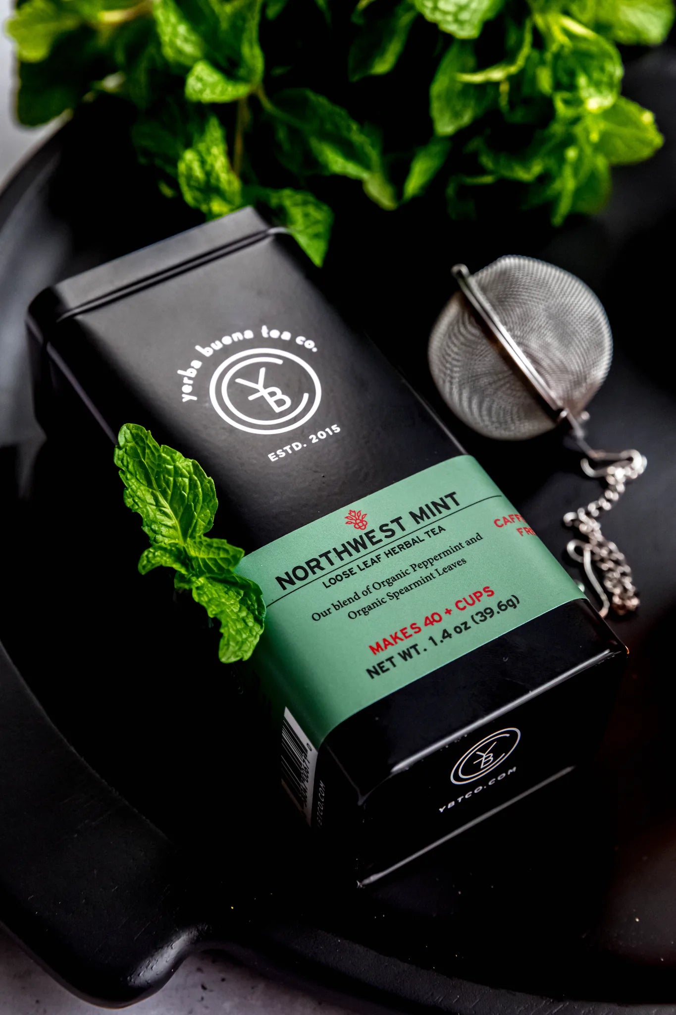Northwest Mint loose leaf herbal tea package displayed on black surface with fresh mint leaves and a metal tea infuser in soft natural light, from Yerba Buena Tea Company.