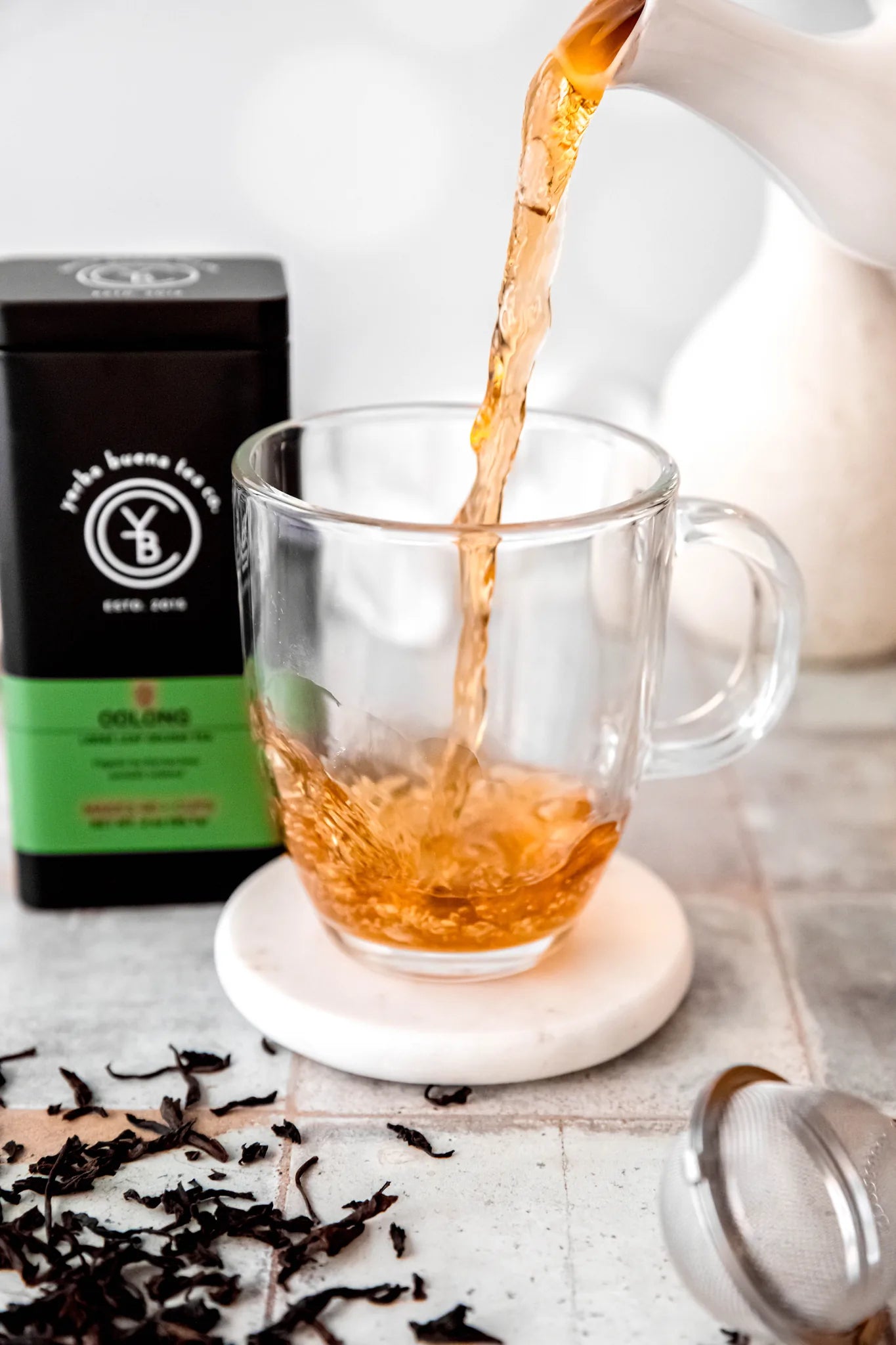 Golden oolong tea being poured into a clear glass mug, with loose dark tea leaves scattered on a marble surface and branded packaging in the background, by Yerba Buena Tea Company.