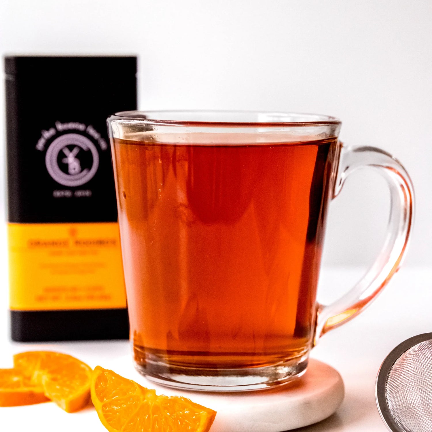 Brewed orange rooibos tea in clear glass mug with fresh orange slices and black packaging, by Yerba Buena Tea Company.