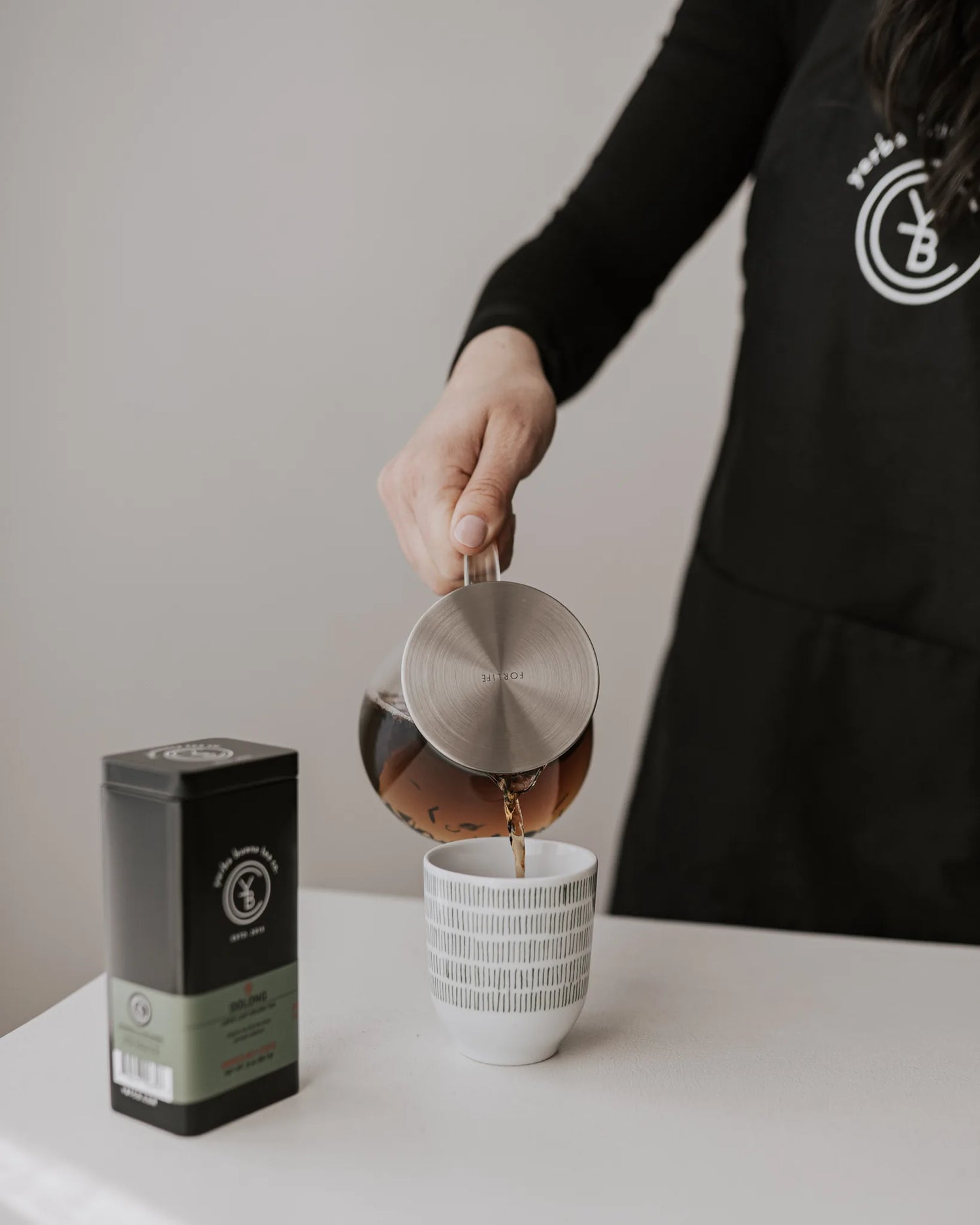 Person in black branded clothing pouring brewed tea from a stainless steel kettle into a white textured ceramic mug, with a black tea canister nearby, by Yerba Buena Tea Company.