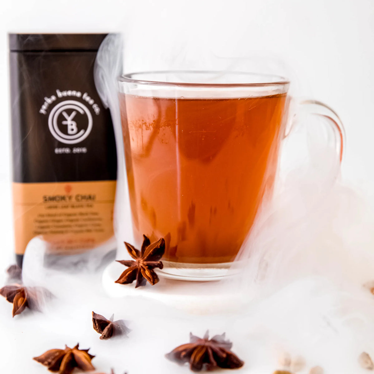 Smoky Chai brewed in a glass mug with wisps of smoke and star anise, its black tin behind, by Yerba Buena Tea Company.