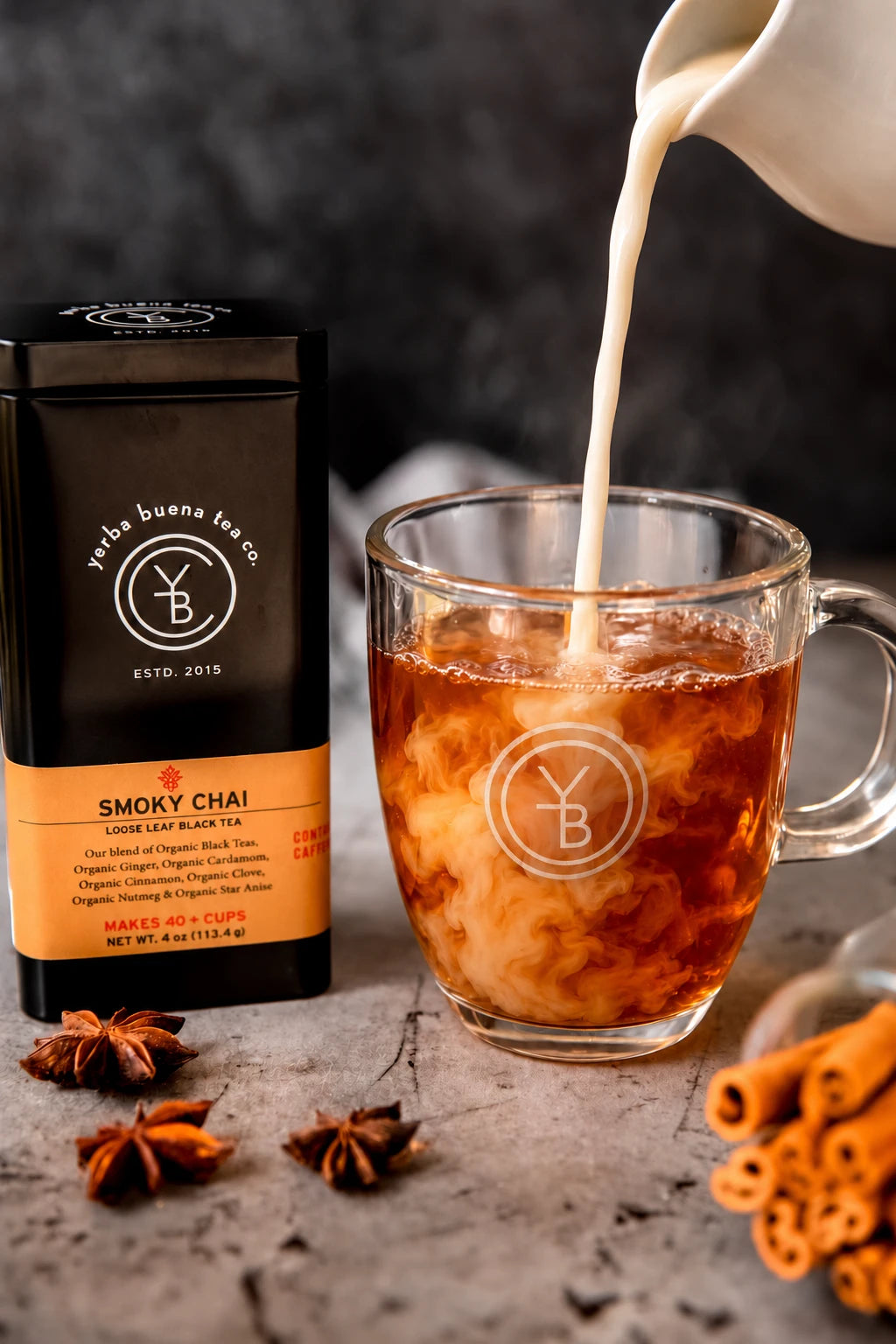 Milk pours into a glass mug of Smoky Chai beside its tin, with star anise and cinnamon sticks