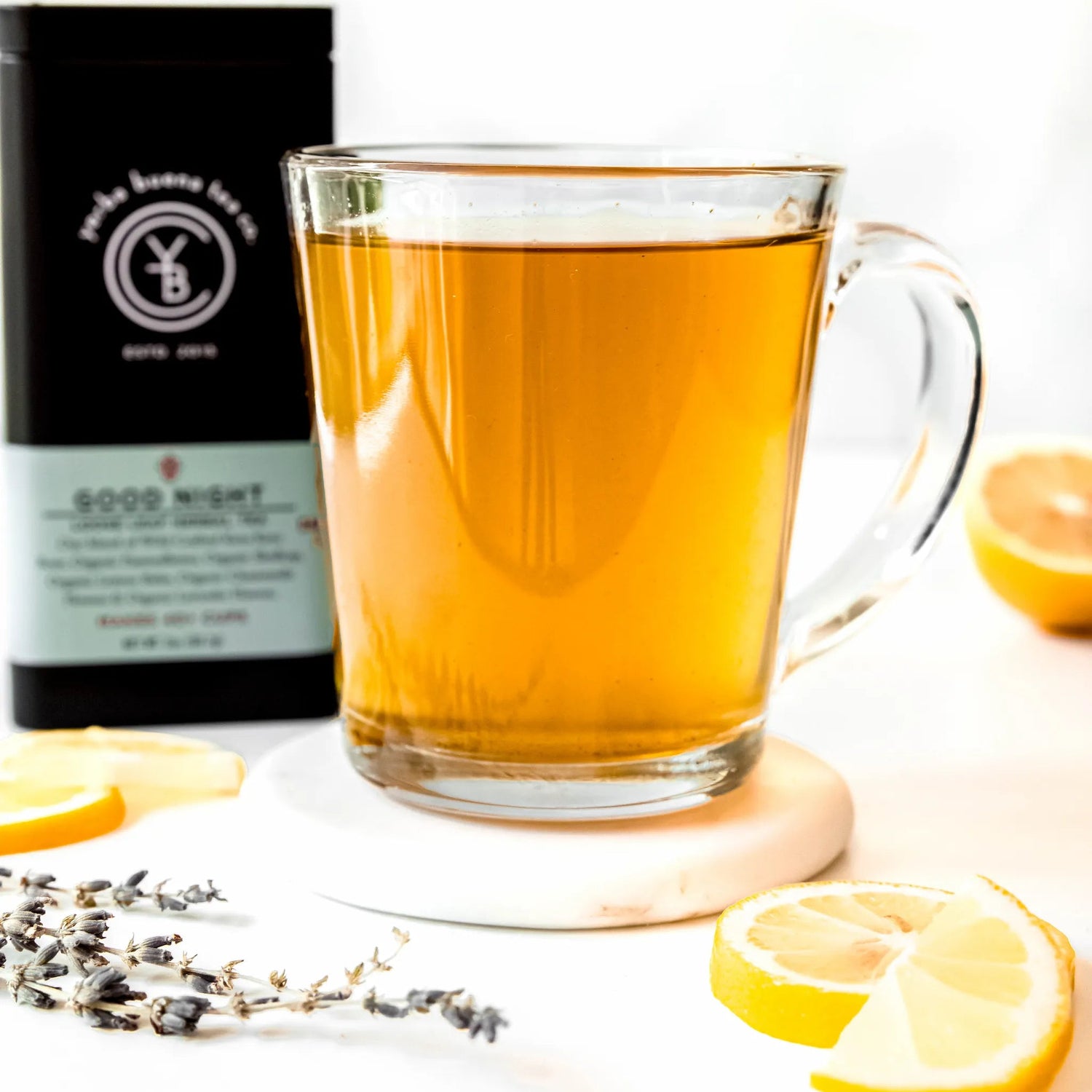 Cup of brewed Good Night herbal tea with lemon slices and lavender, with YBTCO tea tin in the background.