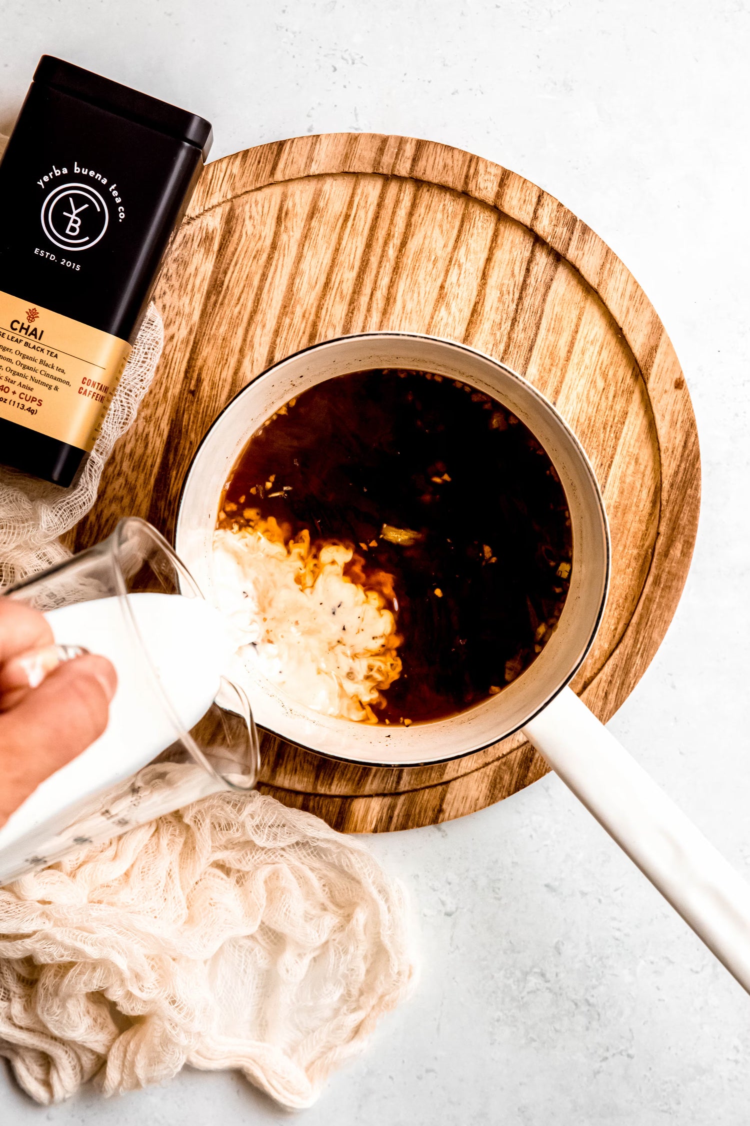 Milk being poured into a saucepan of dark, steeping Chai tea, with the Yerba Buena Tea Co. tin nearby on a rustic wooden board. This moment captures the comforting ritual of preparing a handcrafted, spiced tea.