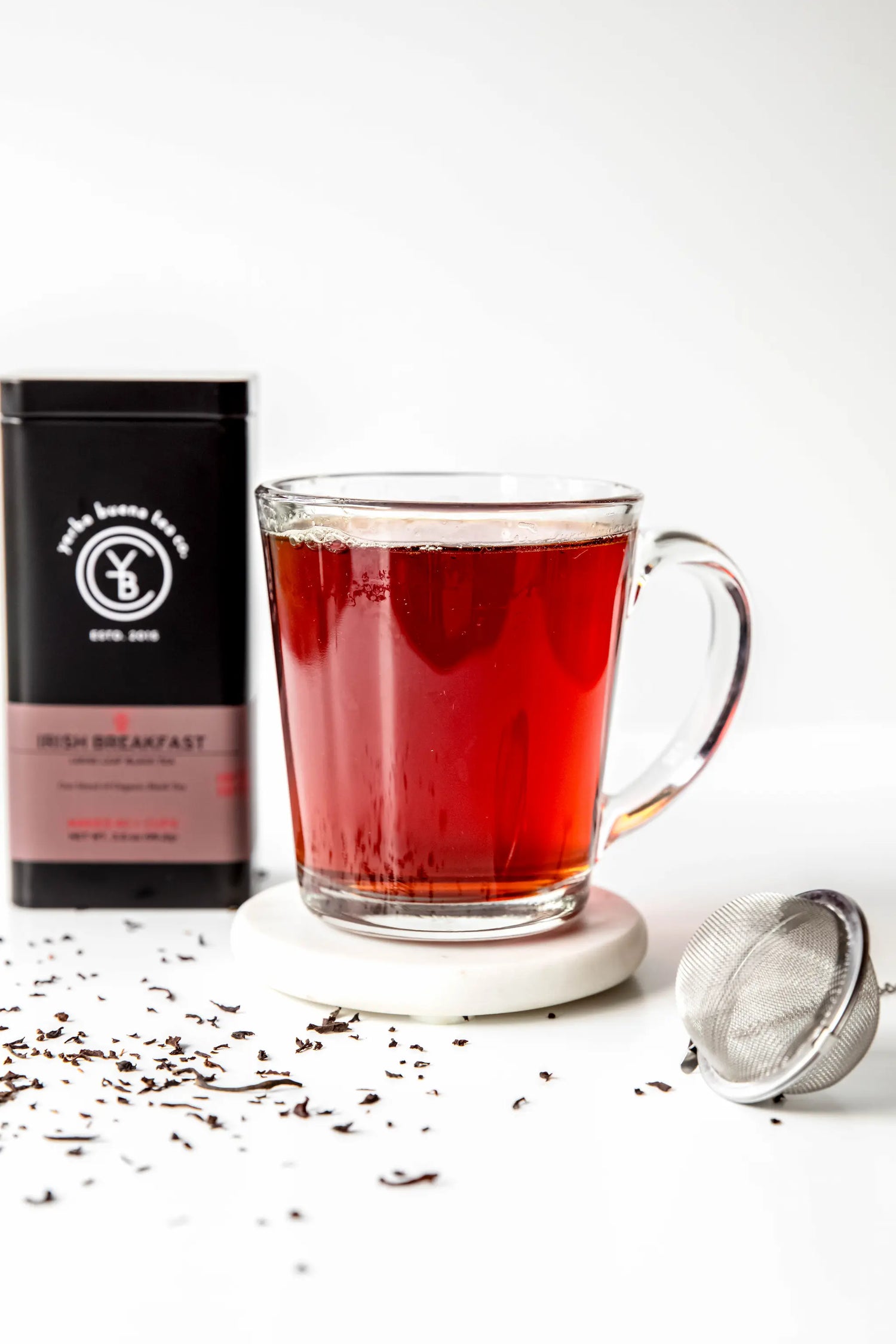 A clear glass mug filled with deeply steeped Irish Breakfast black tea by Yerba Buena Tea Co., styled with scattered loose tea leaves, a metal mesh infuser, and the product tin in the background.