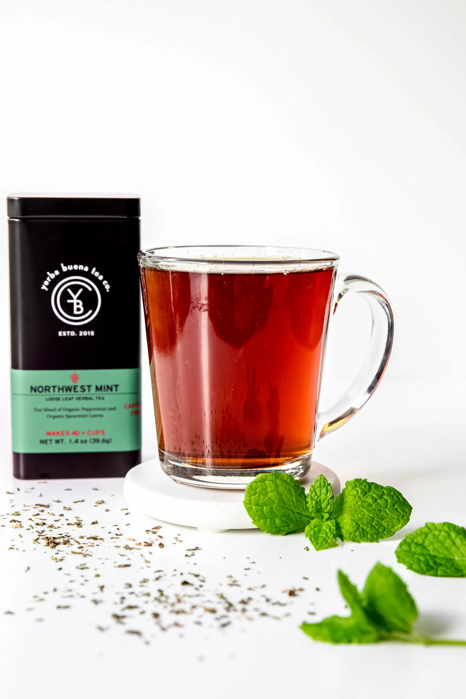 A clear glass mug filled with freshly brewed Northwest Mint herbal tea by Yerba Buena Tea Co., styled with fresh green mint leaves, scattered dried tea, and the product tin in the background.