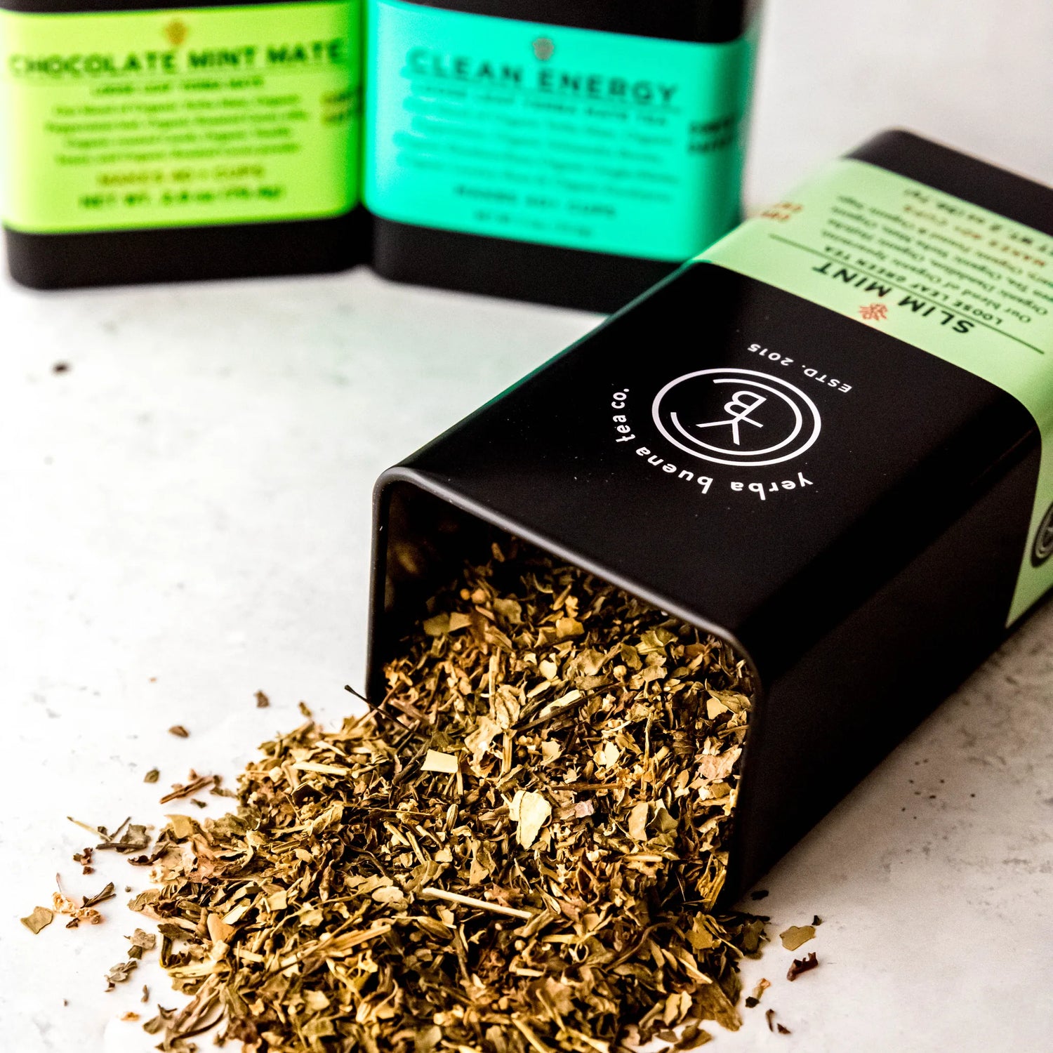 Yerba Mate Box Set with three loose leaf pouches including Chocolate Mint Mate and Clean Energy displayed with spilled tea leaves on marble surface, by Yerba Buena Tea Company.