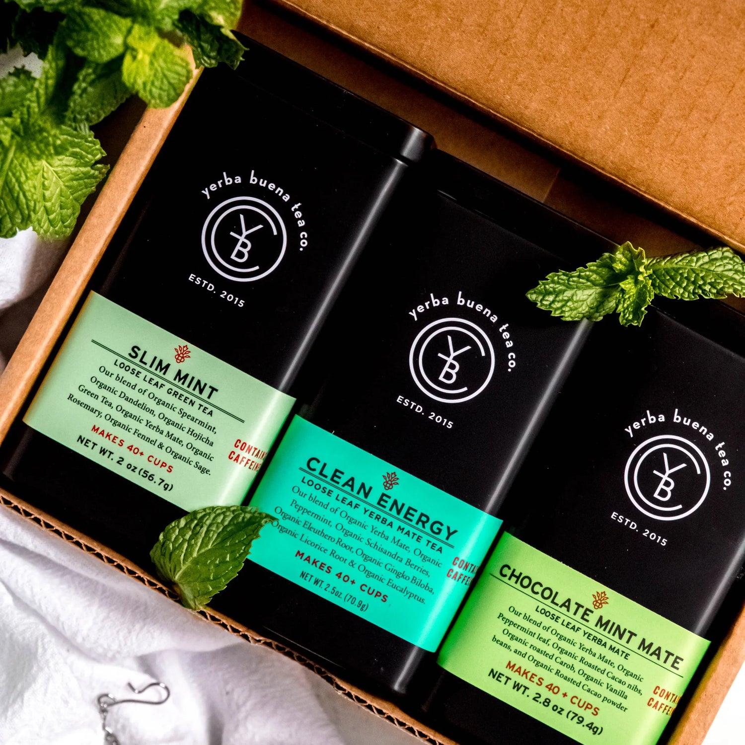 The Yerba Mate gift set containing three loose leaf tea pouches: Slim Mint, Clean Energy, and Chocolate Mint Mate, arranged in a kraft box with fresh mint leaves, by Yerba Buena Tea Company.