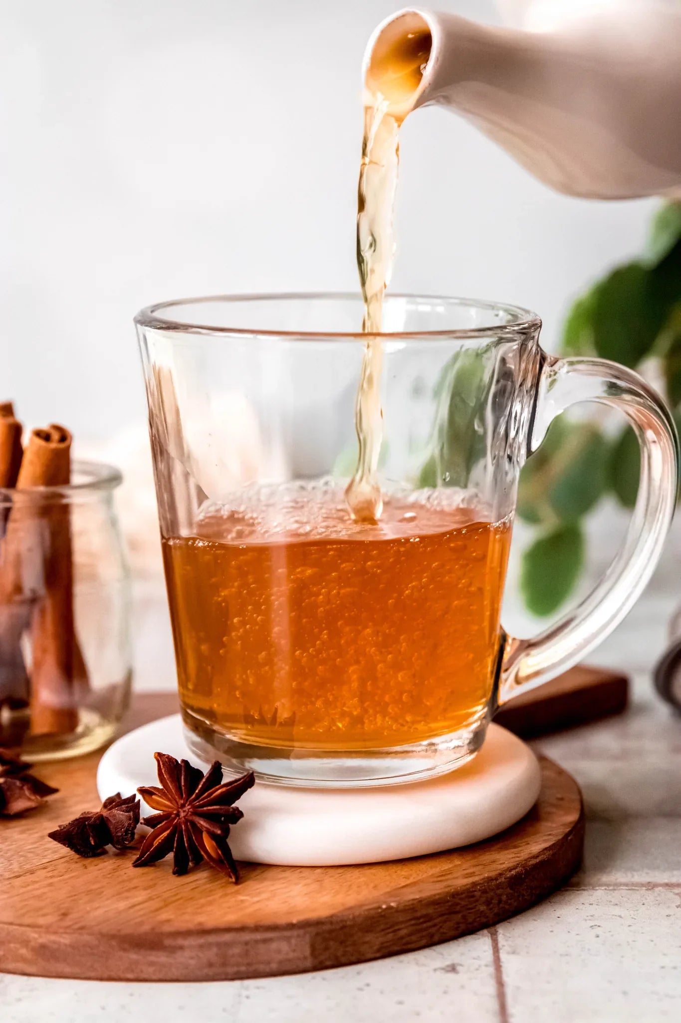 Amber tea pouring from a white ceramic teapot into a glass mug on a wooden coaster with star anise and cinnamon sticks nearby, from Yerba Buena Tea Company.