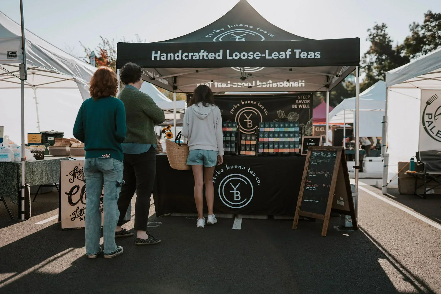 Customers browsing the Yerba Buena Tea Co. booth of organic loose leaf tea tins at an Oregon farmers market in the Willamette Valley.