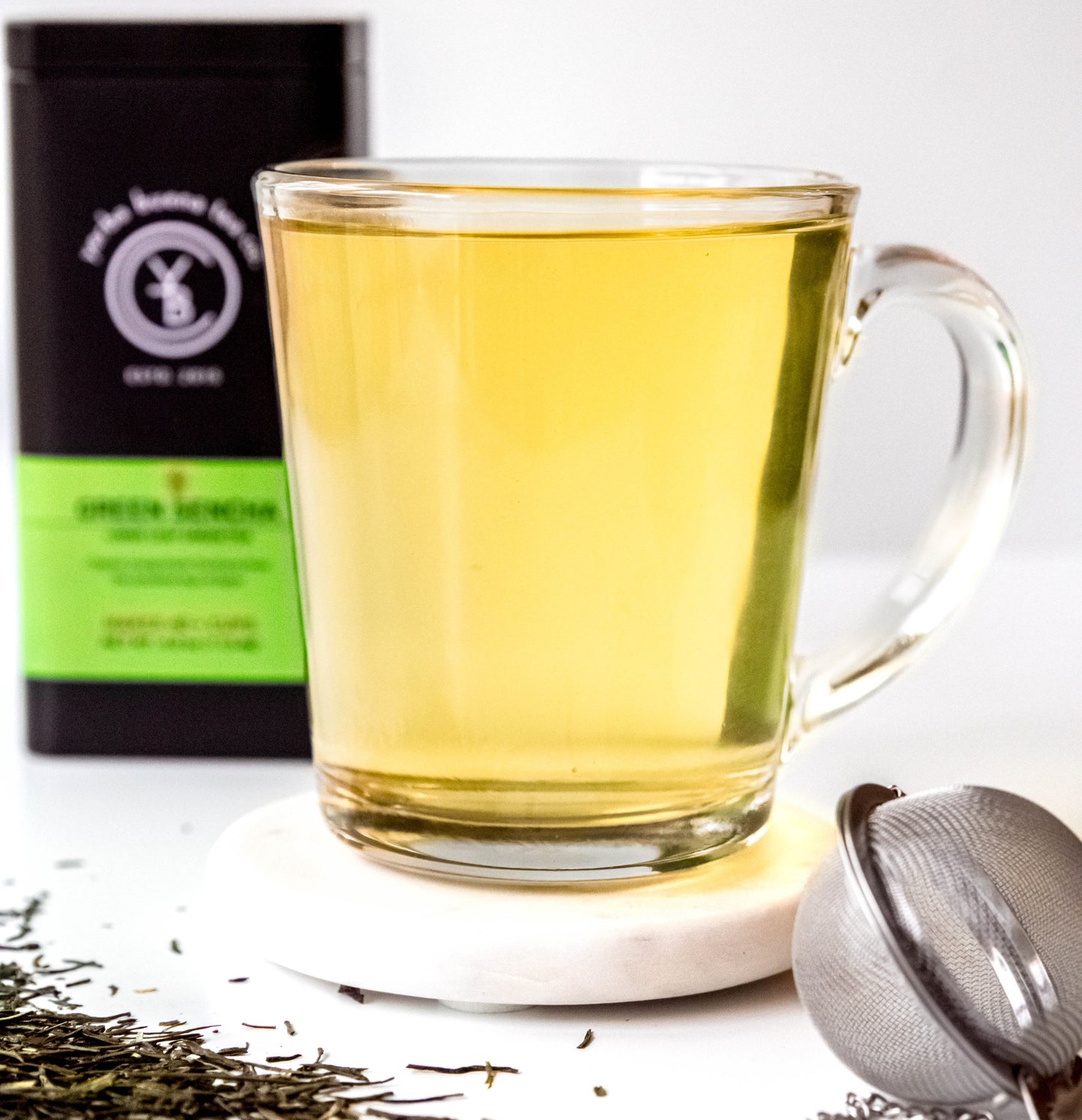 A cup of brewed Green Sencha tea with a light golden-green hue, surrounded by loose-leaf ingredients.