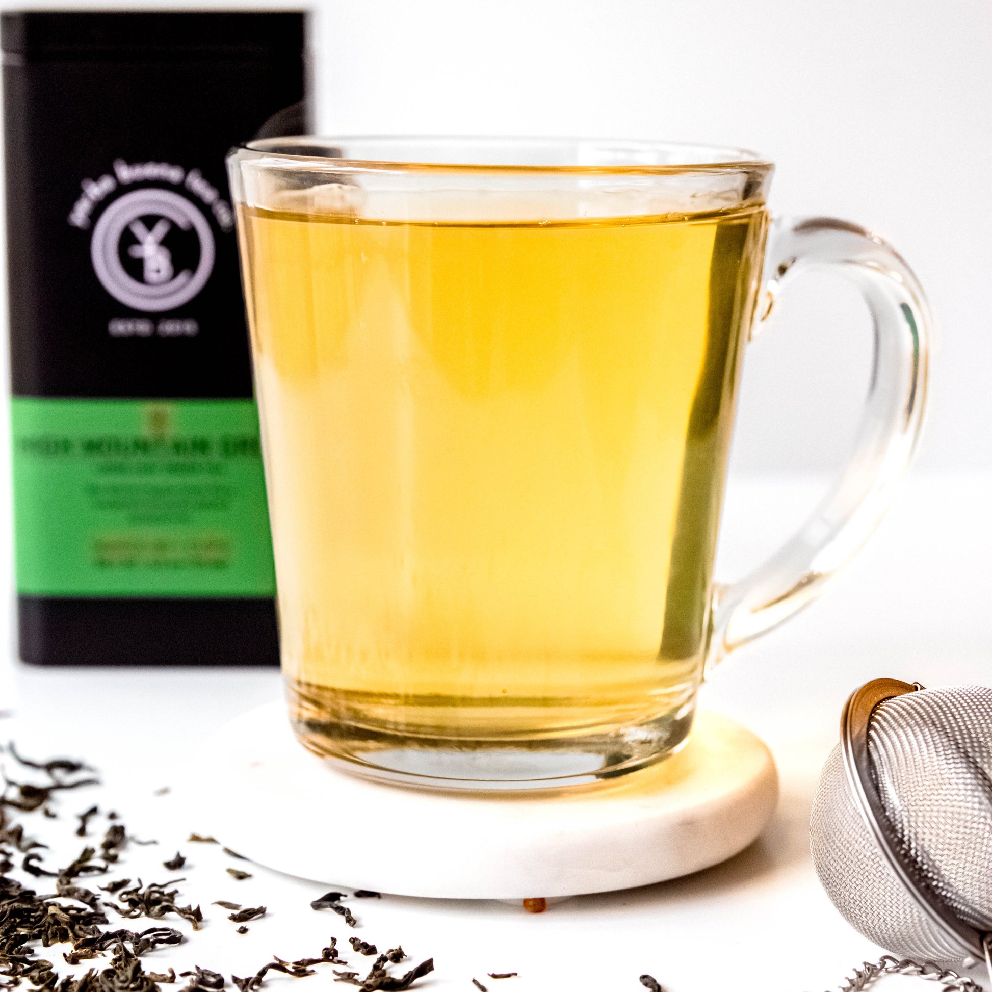 A cup of brewed High Mountain Green tea with a light green hue, surrounded by loose-leaf ingredients.