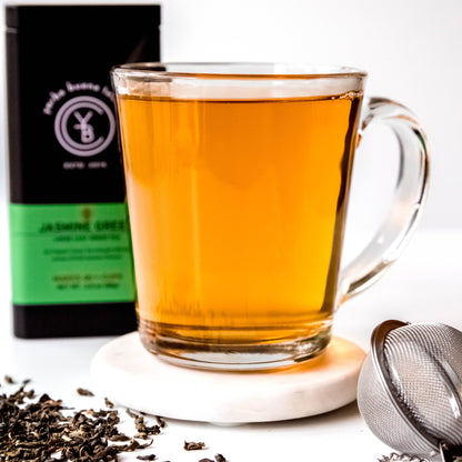 A cup of brewed Jasmine Green tea with a light green hue, surrounded by loose-leaf ingredients.