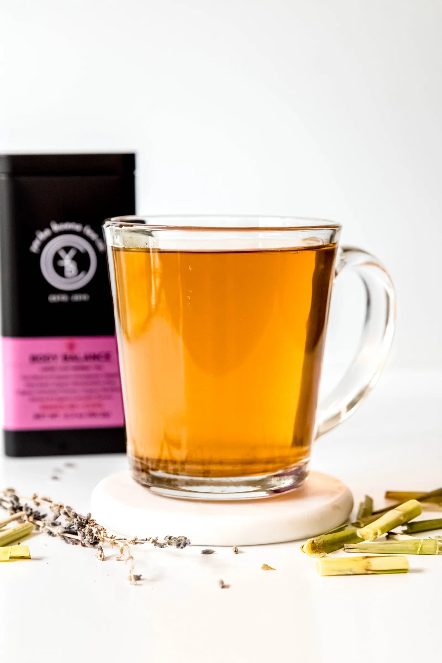 Clear glass mug filled with brewed Body Balance organic herbal tea, light golden in color, resting on a white surface with lemongrass and lavender nearby. A gentle, calming tea ritual handcrafted by Yerba Buena Tea Co.