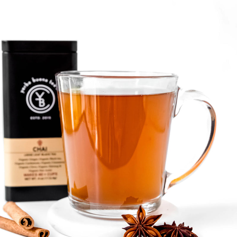 Brewed chai tea in glass mug with cinnamon sticks and star anise beside black canister, by Yerba Buena Tea Company.