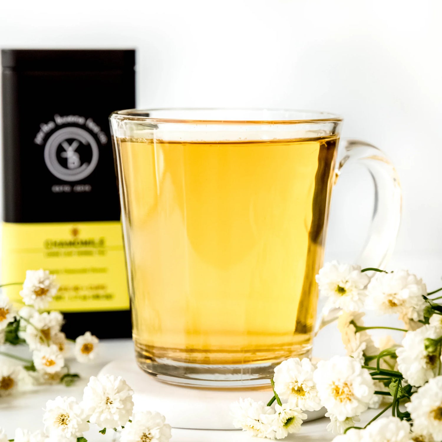 Brewed chamomile tea in clear glass mug surrounded by white chamomile flowers and black packaging pouch, by Yerba Buena Tea Company.