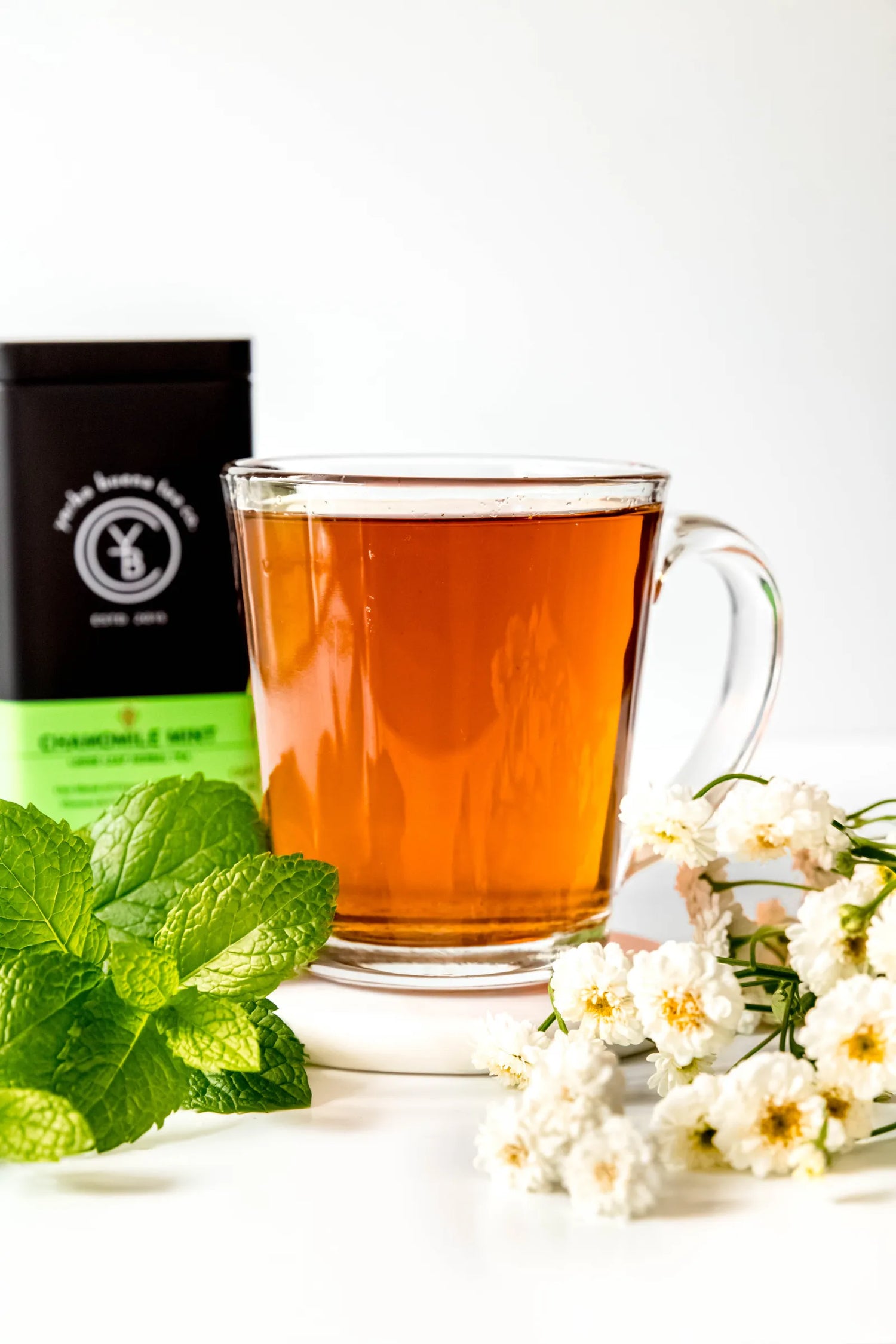 Clear glass mug filled with brewed Chamomile Mint organic herbal tea, golden in color, resting on a white surface with fresh mint leaves and chamomile flowers nearby. A calming, caffeine-free evening tea ritual handcrafted by Yerba Buena Tea Co.