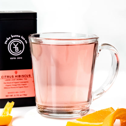 A clear glass cup filled with light pink Citrus Hibiscus tea, placed beside orange slices, with a Yerba Buena Tea Co. tin in the background.