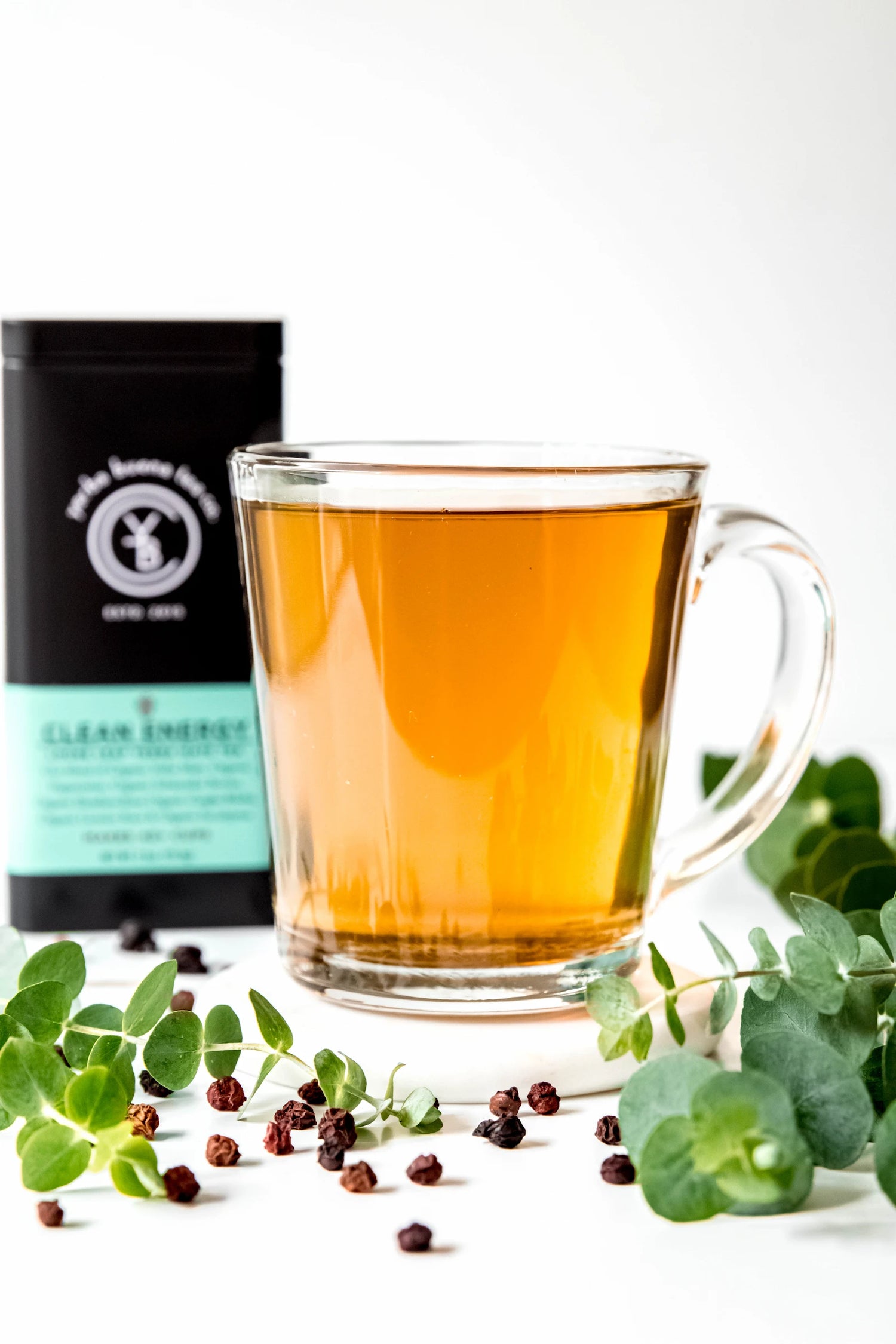 Clear glass mug filled with brewed Clean Energy organic yerba mate tea, light golden in color, resting on a white surface with fresh eucalyptus leaves and whole peppercorns. A clean, focused energy ritual handcrafted by Yerba Buena Tea Co.
