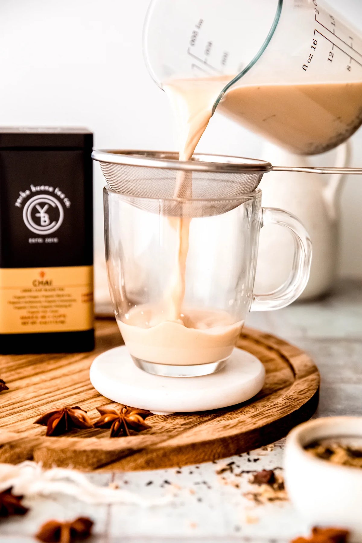 Creamy chai latte being poured through a fine mesh strainer into a clear glass mug, with star anise and loose tea on a wooden board, from Yerba Buena Tea Company.