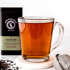 Brewed Darjeeling tea in clear glass mug with loose leaf tea and black pouch packaging visible behind, by Yerba Buena Tea Company.