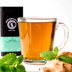 Brewed amber tea in glass mug with fresh mint leaves and ginger root nearby, black and mint packaging behind, by Yerba Buena Tea Company.