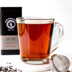 Deep red brewed Earl Grey tea in clear glass mug with infuser ball and black packaging box, by Yerba Buena Tea Company.