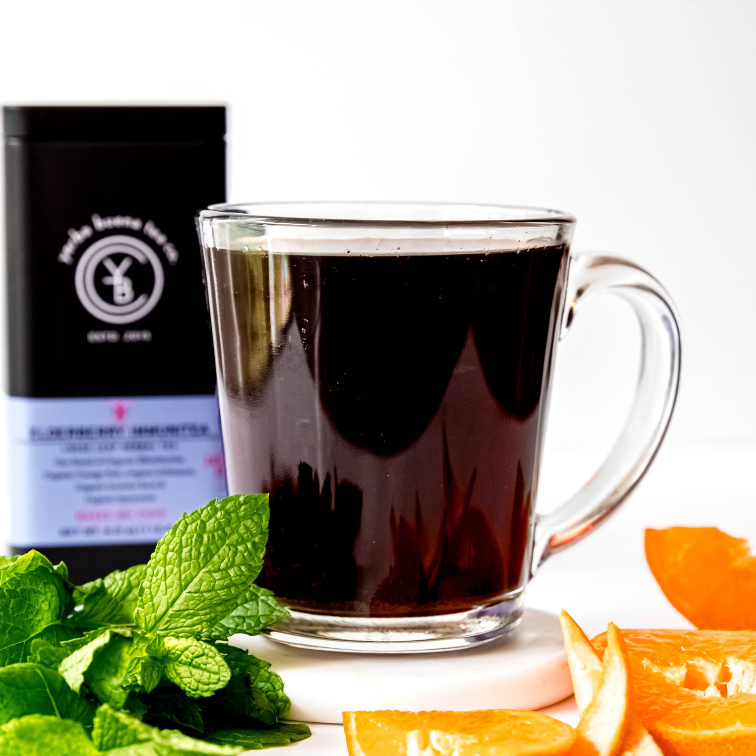 A steaming cup of Elderberry Immunitea in a clear glass mug, with the Yerba Buena Tea Co. Elderberry Immunitea tea tin in the background. Fresh mint leaves and slices of orange are placed beside the cup, highlighting the vibrant and nourishing ingredients of the tea.