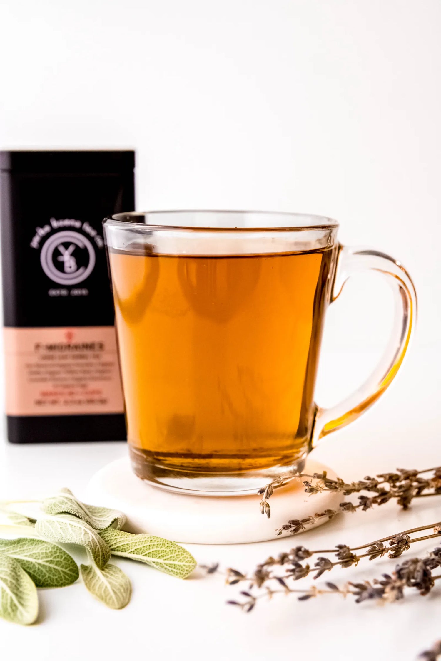 Clear glass mug filled with brewed F-Migraines organic herbal tea, light golden in color, resting on a white surface with sage leaves and lavender sprigs nearby. A calming, herb-forward tea ritual handcrafted by Yerba Buena Tea Co.
