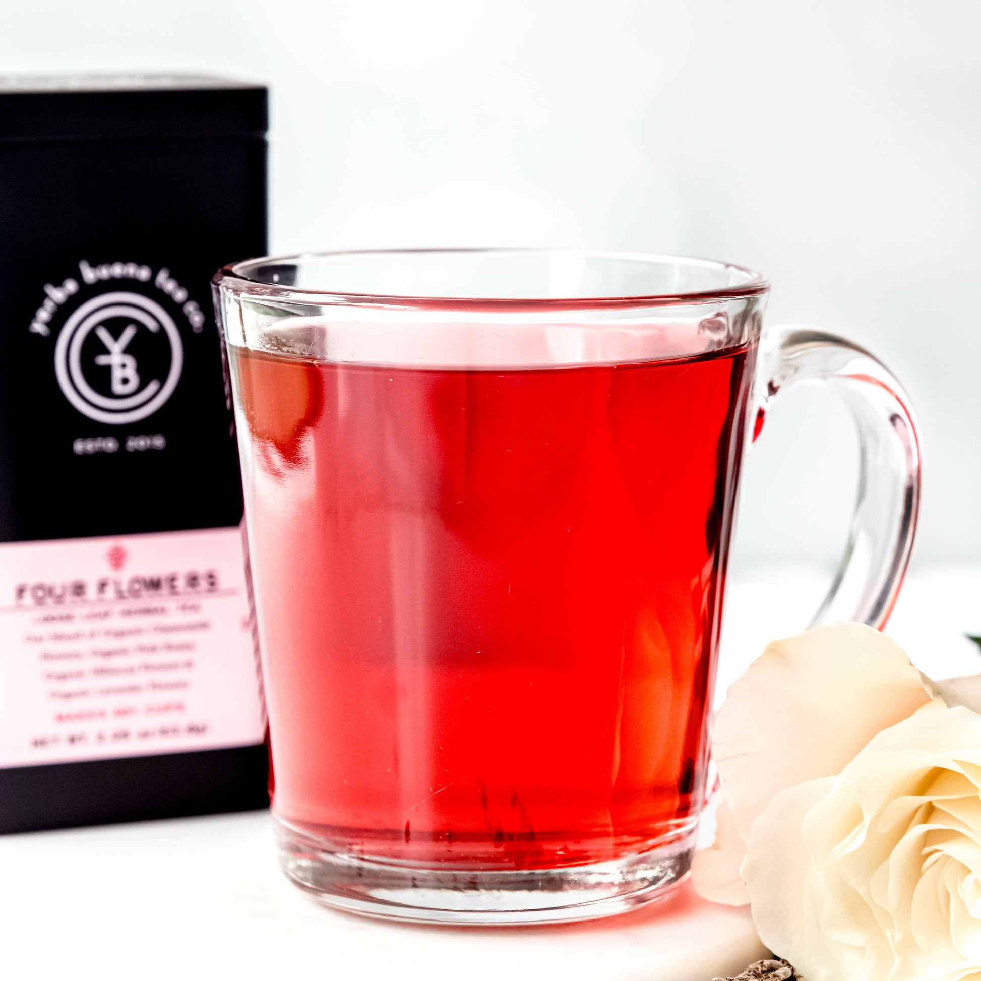 A clear glass cup filled with vibrant red Four Flowers tea, placed beside a white rose, with a Yerba Buena Tea Co. tin in the background.