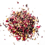 Loose leaf herbal tea blend with red rose petals, chamomile flowers, and dark herbs scattered on white background, by Yerba Buena Tea Company.