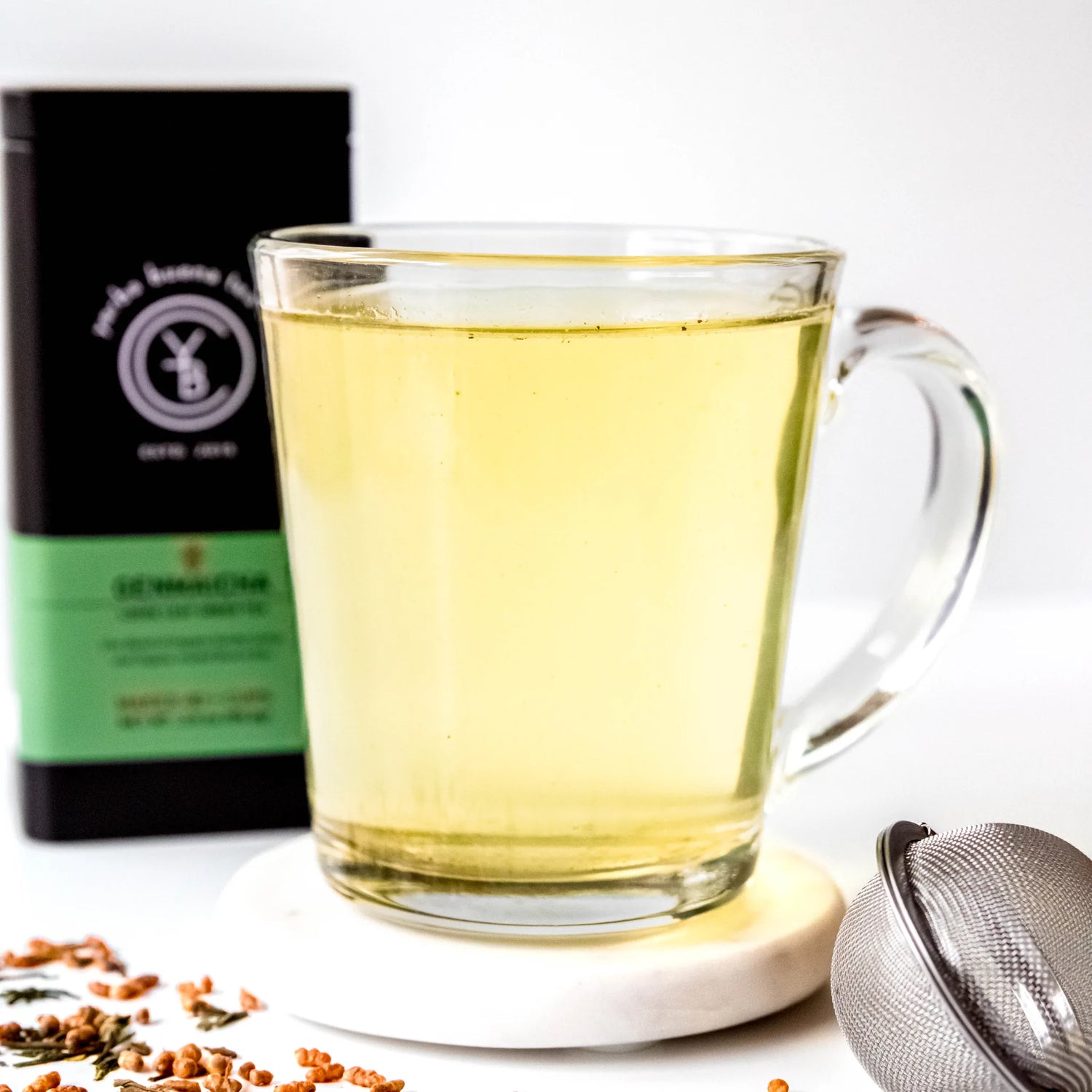 Pale yellow brewed genmaicha tea in a glass mug with infuser ball and packaging pouch behind, by Yerba Buena Tea Company.