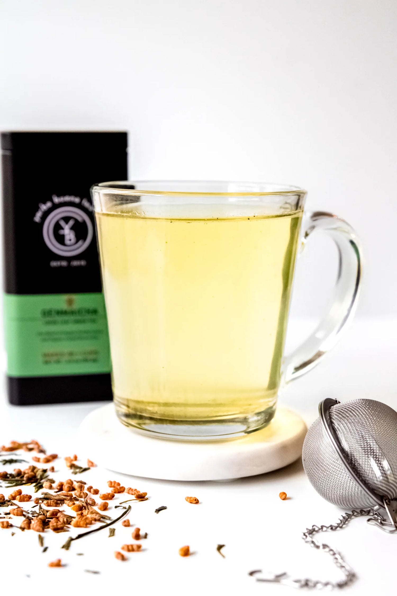 Clear glass mug filled with brewed Genmaicha organic green tea, pale golden in color, resting on a white surface beside toasted brown rice and green tea leaves. A traditional Japanese-style tea ritual handcrafted by Yerba Buena Tea Co.