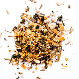 Loose leaf green tea blend with dried citrus pieces and grass-like herbs scattered on white background, by Yerba Buena Tea Company.