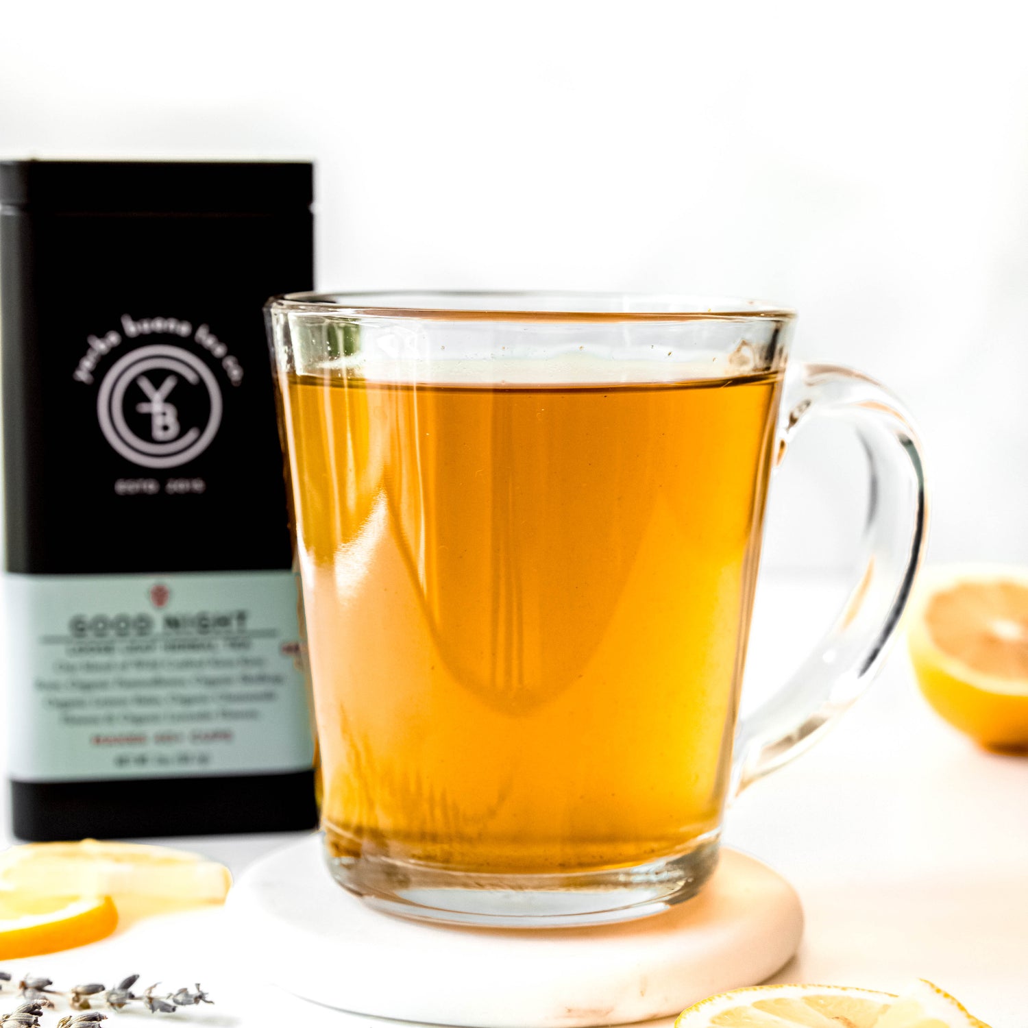 A calming cup of Good Night tea in a clear glass mug, with the Yerba Buena Tea Co. Good Night tea tin in the background, showcasing the light golden-brown hue of the brewed herbal tea. Fresh slices of lemon and sprigs of lavender are placed beside the cup, enhancing the serene and peaceful setting.